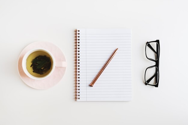 Semplice piatto femminile disteso con tè verde in un taccuino a tazza rosa con penna in metallo e occhiali da lettura