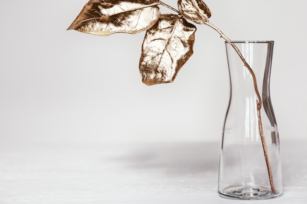 Simple glass vase with branch of tree and bright gold colored leaves on white table. Modern home decoration plant.