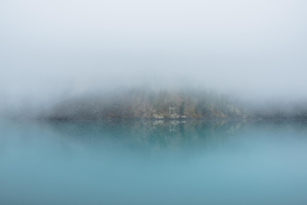 写真 濃霧の中にターコイズブルーの湖があるシンプルな瞑想的な風景 濃霧の中のクリアランスから、色褪せた紅葉と鏡山の湖の急斜面までの静かな景色 反射する紺碧の高山湖