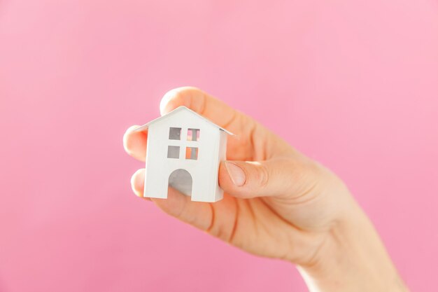 ピンクのパステル調のカラフルなトレンディな背景に分離されたミニチュア白いおもちゃの家を持つ女性の女性の手を単にデザイン