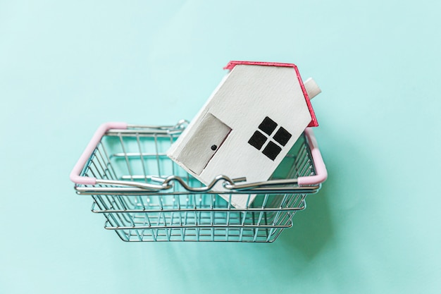 Simply design with miniature white toy house and supermarket grocery shopping basket isolated on blue background.