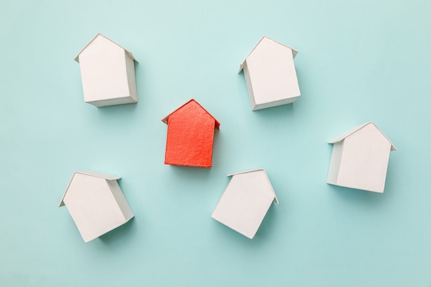 Simply flat lay design with miniature red toy model house in\
among white houses isolated on pastel blue background. real estate\
property industry. community unique neighborhood choice\
concept.