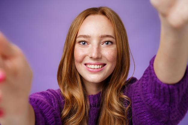 Photo sincere bright and cheerful ginger girl in purple sweater reaching camera with hands and smiling broadly with friendly optimistic gaze, taking selfie or wanting hold something over violet wall.