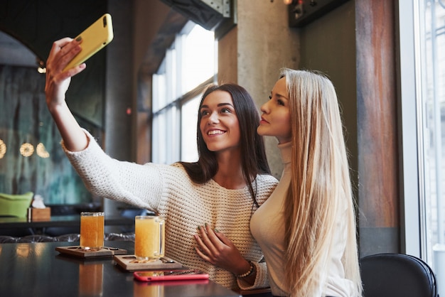 Sincere brunette has nice smile. Young female friends take selfie in the restaurant with two yellow drink on table