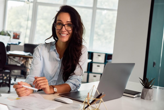 A sincere business woman in glasses is working on a laptop in the office and smiling at the camera