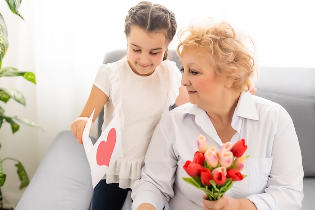 Sincere cute little granddaughter strong cuddling and kissing in cheek her 60s grandmother gave her pretty spring flowers congratulates with birthday, International Womens day, close up concept image.