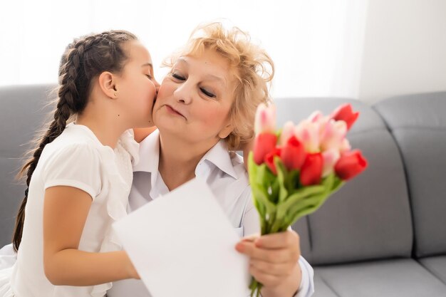 Sincere cute little granddaughter strong cuddling and kissing in cheek her 60s grandmother gave her pretty spring flowers congratulates with birthday, International Womens day, close up concept image.