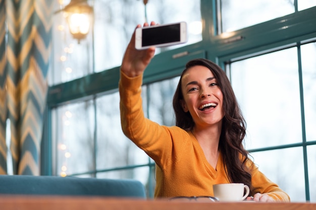 Sincere selfie. Beautiful merry cute woman laughing while elevating phone and taking shot