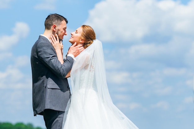 Sincere smiles of the newlyweds hugging on the background of the blue sky