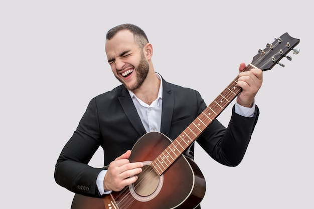 Singer in black suit stand and play on guitar. He sing song. Young man wears suit. 