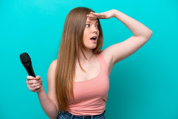 Singer caucasian woman picking up a microphone isolated on blue background doing surprise gesture while looking to the side