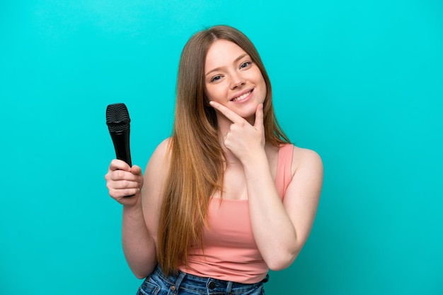 Singer caucasian woman picking up a microphone isolated on blue background happy and smiling