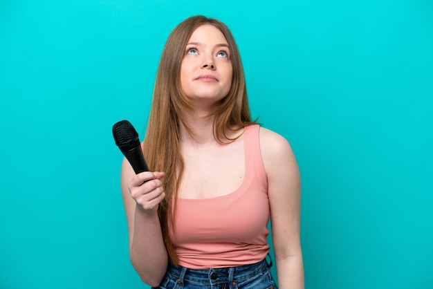 Singer caucasian woman picking up a microphone isolated on blue background and looking up