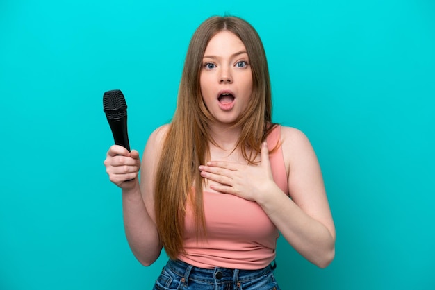Singer caucasian woman picking up a microphone isolated on blue background surprised and shocked while looking right