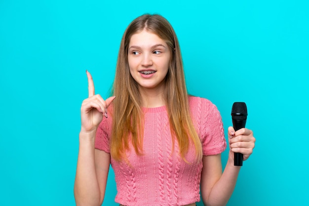 Singer Russian girl picking up a microphone isolated on blue background intending to realizes the solution while lifting a finger up