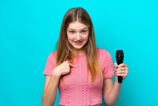 Singer Russian girl picking up a microphone isolated on blue background with surprise facial expression