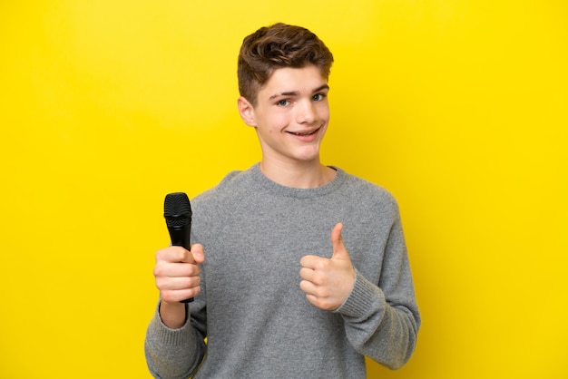 Singer Teenager man picking up a microphone isolated on yellow background with thumbs up because something good has happened