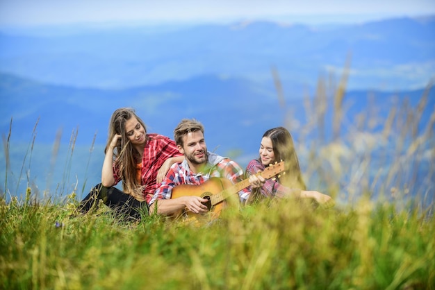 一緒に歌うミュージカルポーズハイキングエンターテインメント静かな場所自然のメロディーハイキングの伝統音楽でハイキングする友達山の頂上でリラックスしながらハンサムな男がギターを弾く