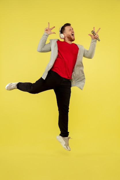 Singing with headphones. Full length portrait of young high jumping man isolated on yellow studio background. Attractive male caucasian model. Copyspace. Human emotions, facial expression concept.