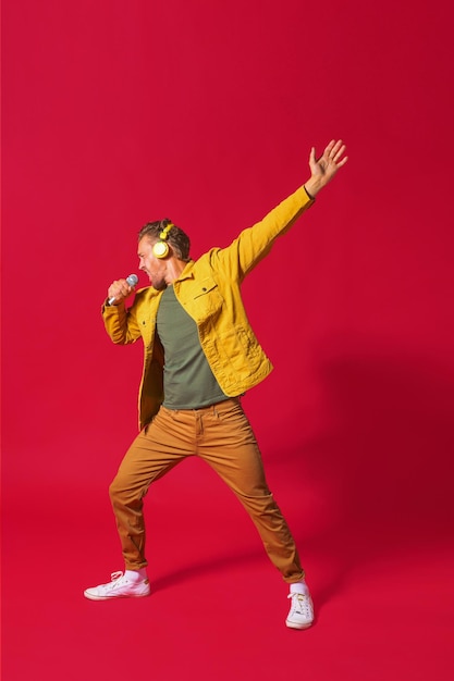 Фото Поющий молодой человек наслаждается песней и танцует с поднятой рукой, используя телефон и беспроводные наушники в джинсовой желтой куртке на красном фоне мужчина поет, записывая свой голос