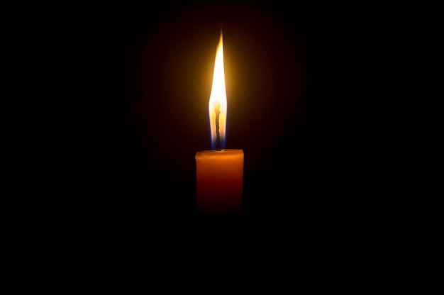 A single burning candle flame or light glowing on an orange candle on black or dark background on table in church for Christmas funeral or memorial service with copy space