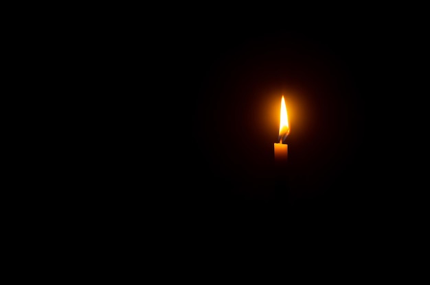 A single burning candle flame or light glowing on an orange candle on black or dark background on table in church for Christmas funeral or memorial service with copy space