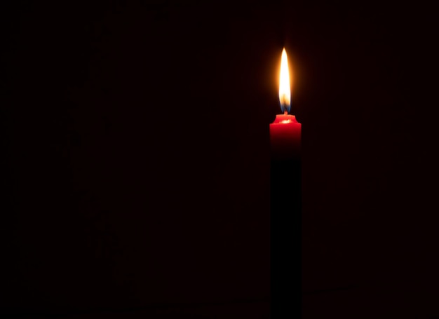 Single burning candle flame or light glowing on a small red candle on black or dark background