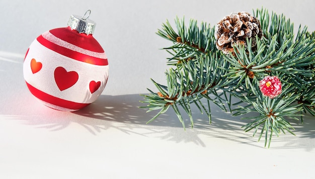 Single Christmas decorative trinket Red white glass ball with heart shapes Fir twig with cone Sunlight with long shadows on off white paper Festive Xmas design Panoramic banner composition