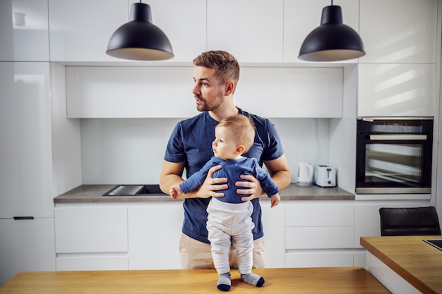Single dad standing in kitchen with his beloved son. Toddler standing at kitchen table and father holding him.