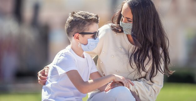 Single mom is looking worried at her son in sunglasses outside, both wearing face masks.