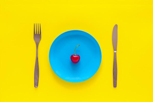 Singola ciliegia matura sul piatto blu, coltello, forchetta su sfondo giallo