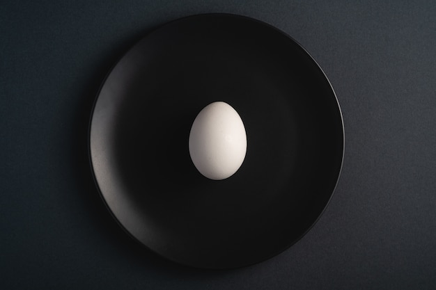 Single white egg in black plate on dark moody plain minimal surface, top view, happy Easter day