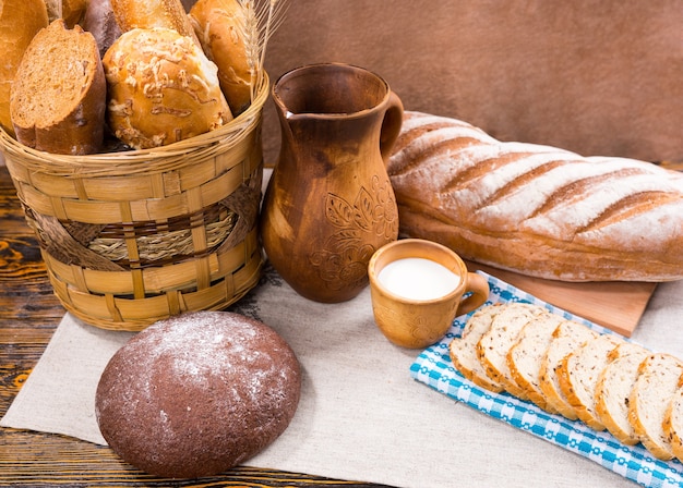 Single wooden pitcher, full cup of milk and freshly baked bread as whole loaf and slices next to basket on table