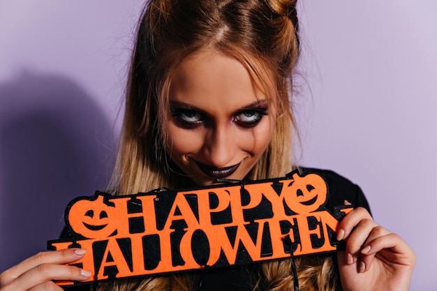 Sinister ragazza dai capelli biondi che guarda alla fotocamera con un sorriso felice giovane donna con un trucco scuro spaventoso in posa su sfondo viola in halloween