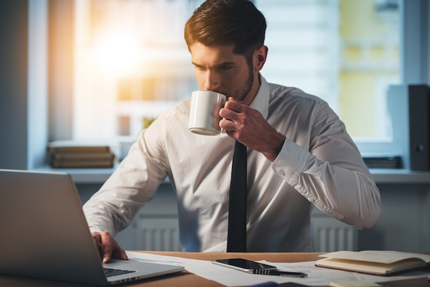 仕事で淹れたてのコーヒーを一口。彼のラップトップを使用してコーヒーを飲む物思いにふける若いハンサムな男