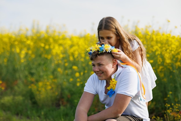 Sister braids ribbons in Ukrainian wreath with flowers on older brother's head on meadow against field