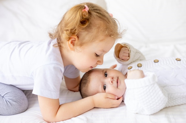 Photo sister gently hugs and kisses a newborn baby in a crib on a white insulated cotton bed two children in the family brother and sister