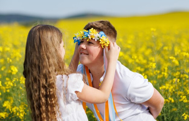 Sister puts on brother Ukrainian wreath with ribbons against background of fields and sky