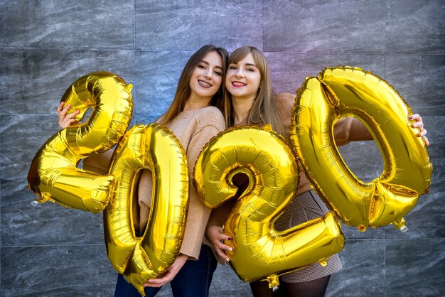 Sisters celebrating new 2020 year. They holding golden air balloons at party celebration