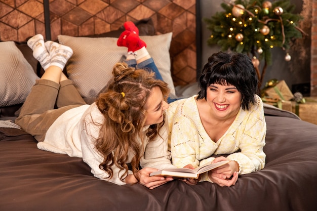 Sisters lying on the bed and reading book. They laughing and celebrating New year eve and Christmas. There are gifts and decorated fir branches with golden balls.