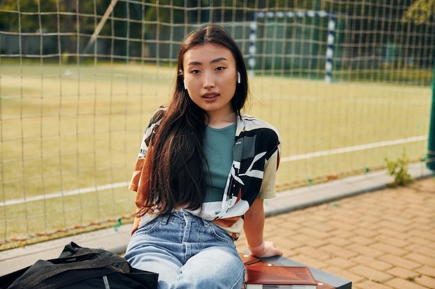 Sits against sport field Young asian woman is outdoors at daytime