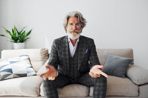 Sits on comfortable sofa in formal clothes Senior stylish modern man with grey hair and beard indoors