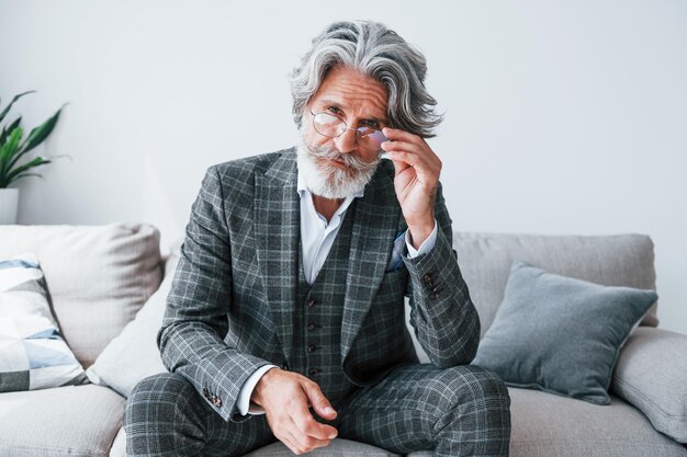 Сидит на удобном диване в формальной одежде Старший стильный современный мужчина с седыми волосами и бородой в помещении