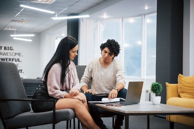 Sits on a grey chairs. Curly guy and brunette girl discusses the details of the contract in modern office