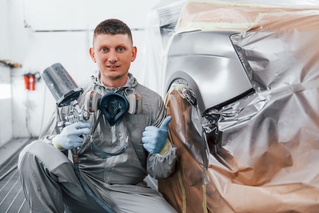 Sits near freshly painted car Caucasian automobile repairman in uniform works in garage