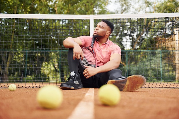 Sits near net and taking a break African american man in pink shirt sits with tennis racket on the court outdoors