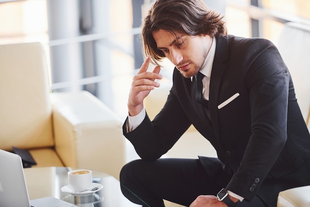 Sits on the sofa. Portrait of handsome young businessman in black suit and tie.