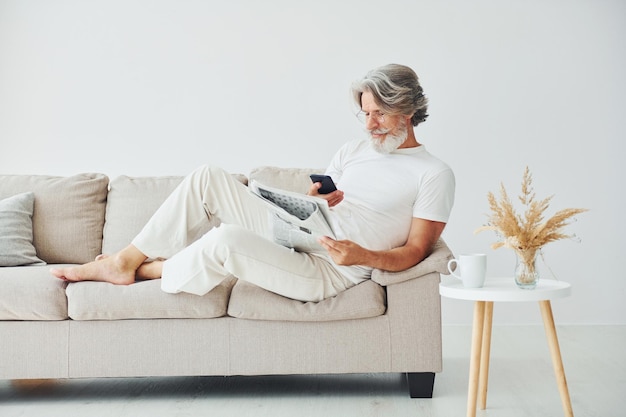 Sits on the sofa Senior stylish modern man with grey hair and beard indoors