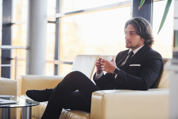 Sits on the sofa with cup of drink. Portrait of handsome young businessman in black suit and tie.