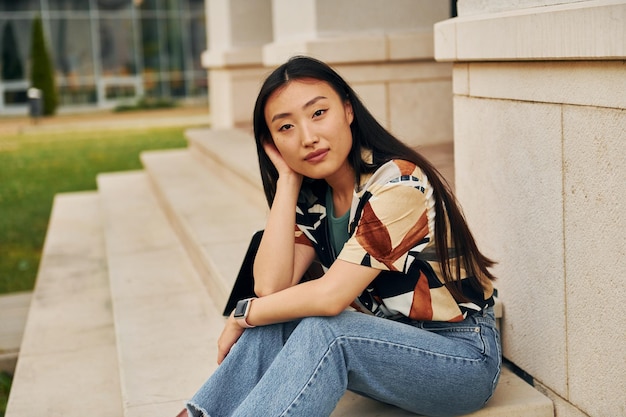 Sits on the stairs of building Young asian woman is outdoors at daytime